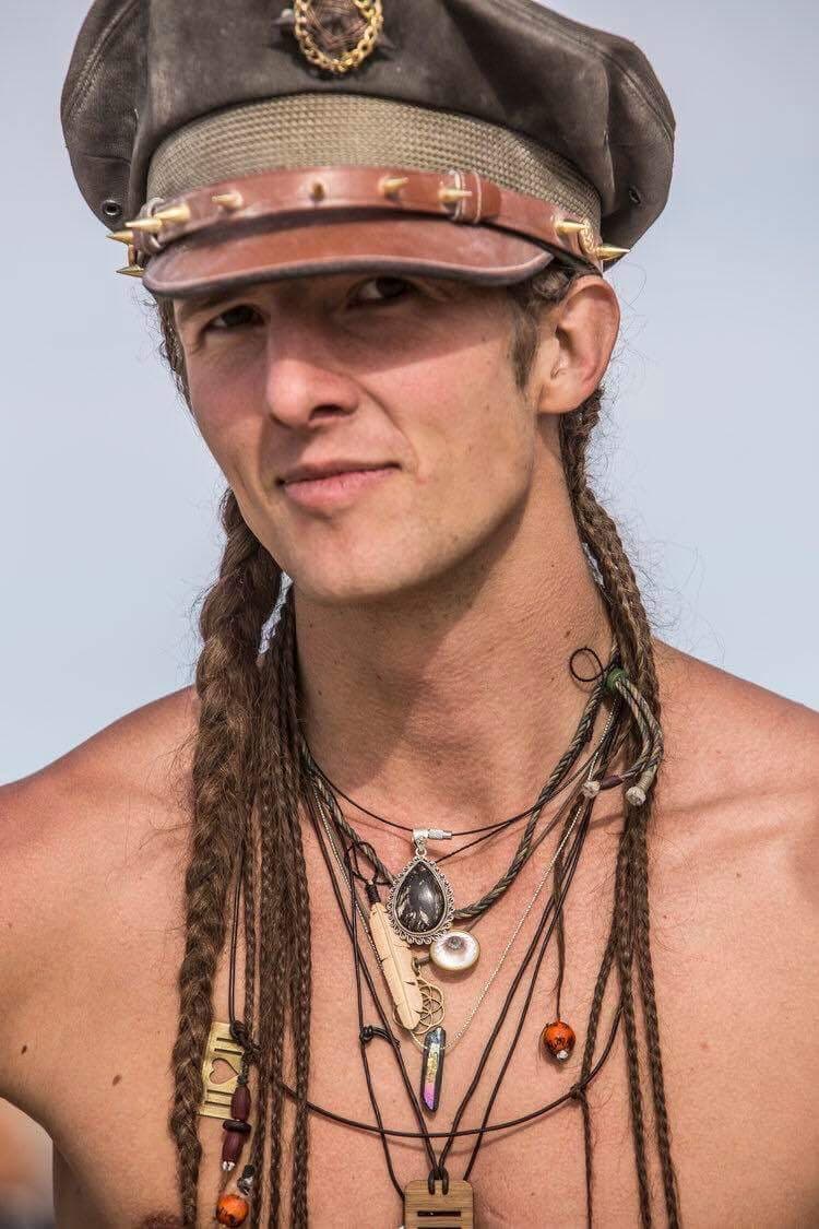 The image features a person wearing a decorative hat and multiple necklaces. The individual has braided hair and is looking directly at the camera with a slight smile.