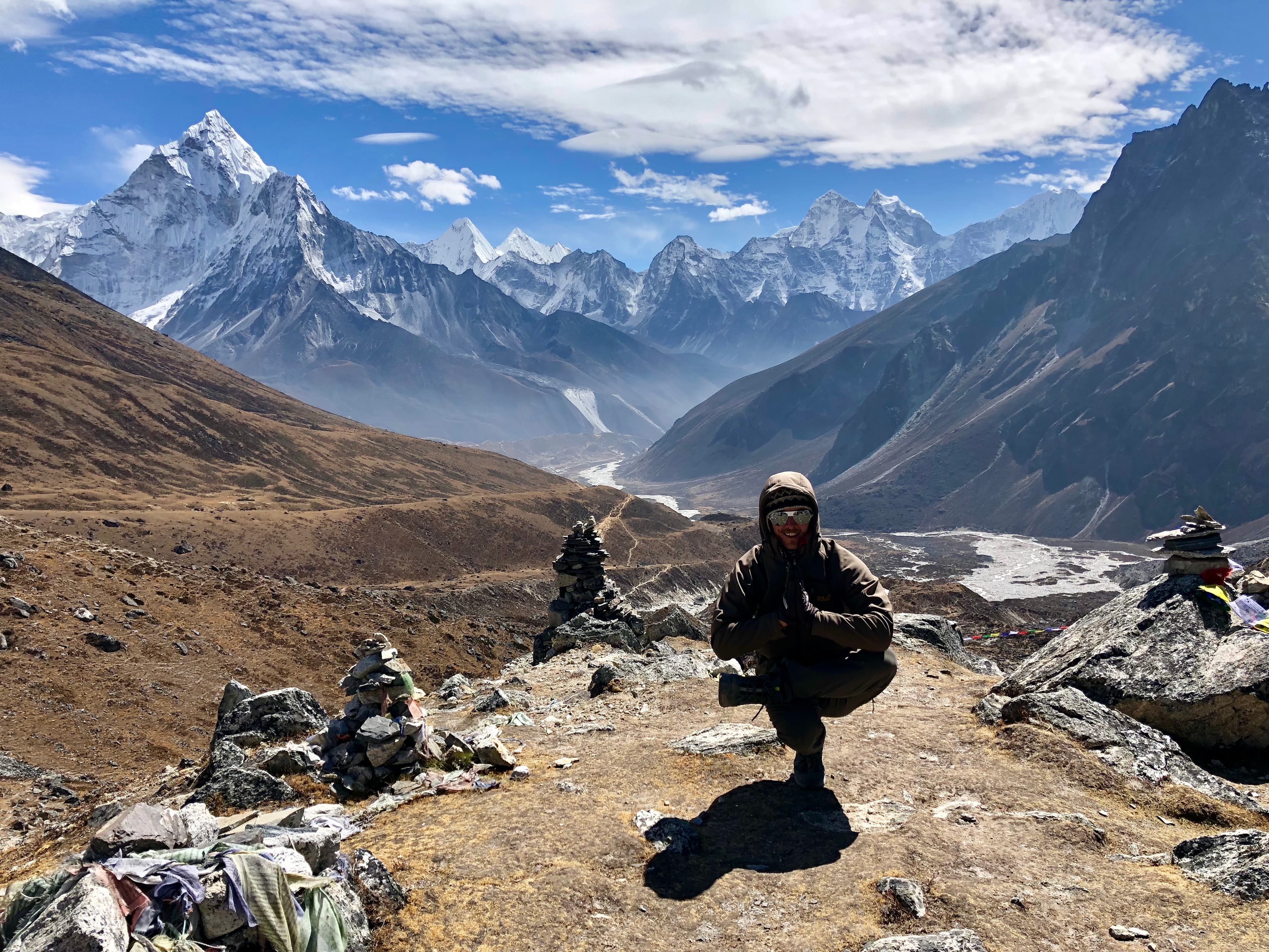 A person is crouching on a rocky mountain path with a stunning backdrop of snow-capped mountains under a partly cloudy sky.