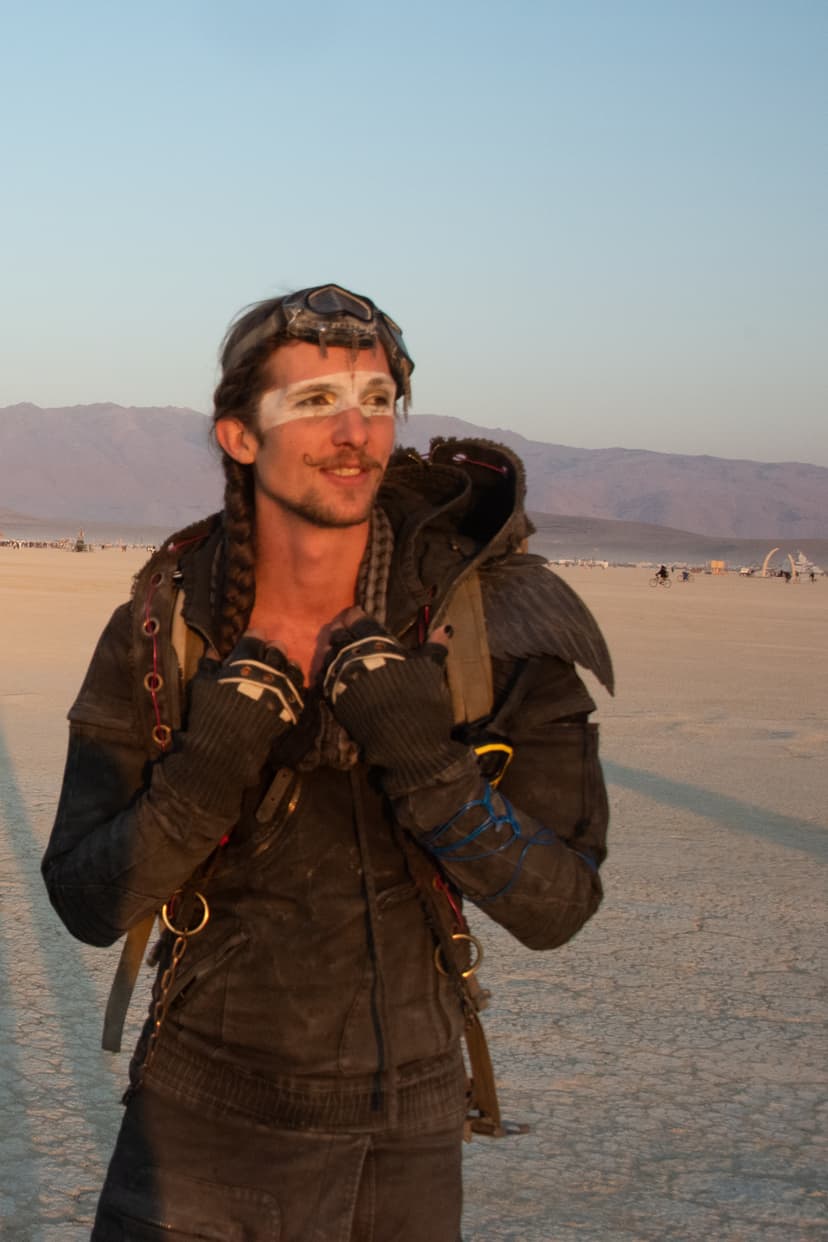 The image shows a person standing outdoors in a desert-like environment. The individual is wearing a costume with a post-apocalyptic style, including goggles on their head, braided hair, and face paint. The background features a vast open space with mountains in the distance.