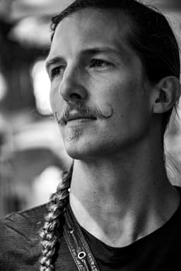 A black and white portrait of a person with long hair tied in a braid and a distinctive mustache, looking thoughtfully into the distance.